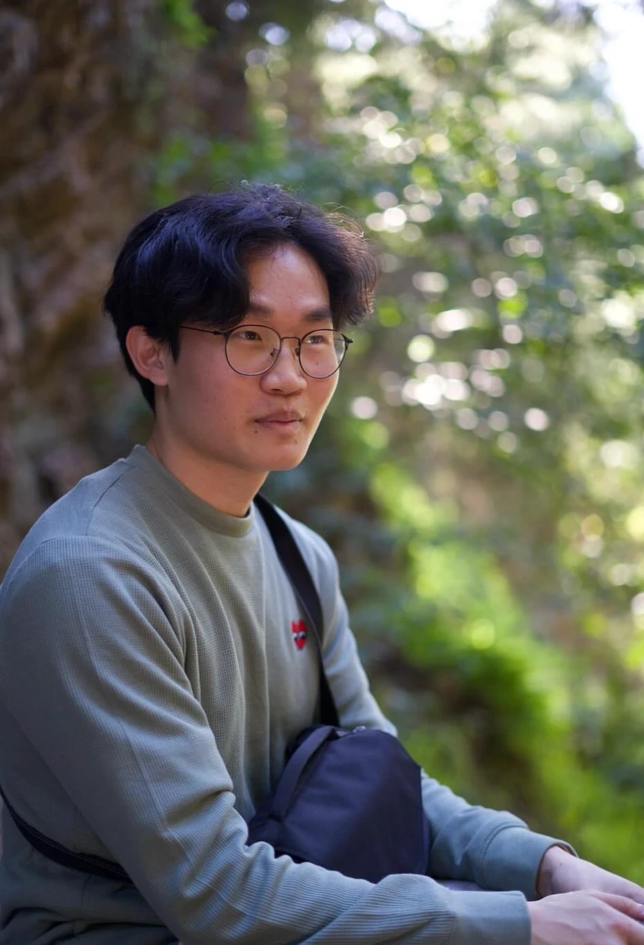 Gordon sitting outdoors among trees, wearing glasses and a green sweater